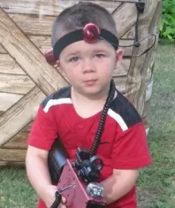 A young boy holding a laser tag tagger in front of a barrier that looks like a wooden fence.