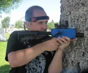 A man holding a blue laser tag tagger in front of a tree.